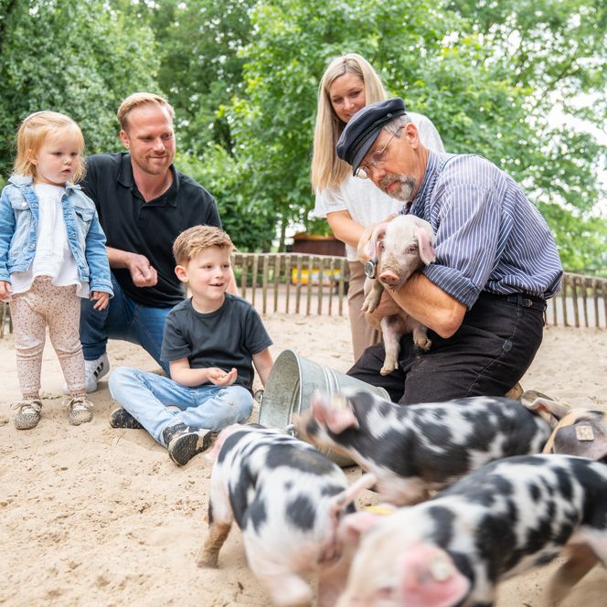 Eine Familie mit Vater, Mutter und zwei kleinen Kindern zwischen 3 und 6 Jahren stehen in einem Schweinegehege und betrachten einen Mann in traditioneller Kleidung, wie er ein Ferkel auf dem Arm hält