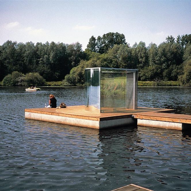 Auf einem See ist ein Holzsteg zu sehen, auf dem sich ein dreieckiger Pavillon aus Glas befindet