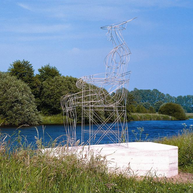 Ein Kunstwerk aus Draht steht auf einem Betonfundament. Im Hintergrund ein Fluss, gesäumt von grünen Bäumen und Pflanzen